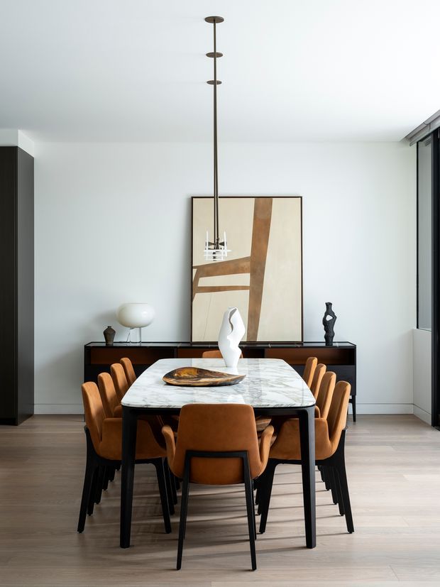 A refined dining room with a long rectangular marble dining table surrounded by caramel-brown upholstered chairs. A dark timber sideboard spans the rear wall, styled with sculptural objects, a white table lamp, and abstract artwork. The room has pale timber flooring, white walls and ceiling, slim dark-framed glazing, and a linear pendant light suspended above the table.