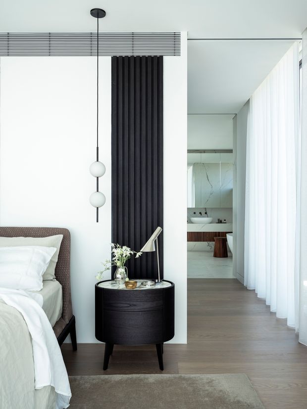 A refined contemporary bedroom with a partially visible upholstered bed, white bedding, dark rounded bedside table, black vertical feature panelling, and a sculptural pendant light. Floor-to-ceiling sheer curtains line the right side, opening toward a bright ensuite bathroom visible in the background. The palette is predominantly white, light timber, grey, and black, with clean minimalist detailing.