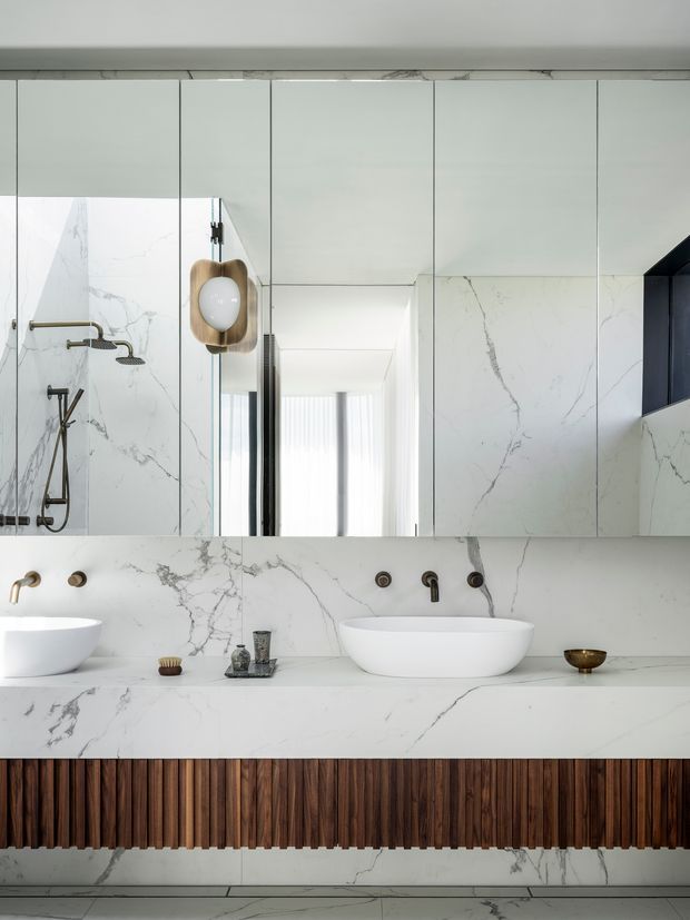 A refined contemporary bathroom featuring a double vanity with sculptural white vessel basins, a freestanding white bathtub, brushed brass and dark tapware, and extensive white marble-look wall and countertop surfaces with prominent grey veining. A mirrored wall visually expands the room, while a warm fluted dark timber vanity front adds texture and contrast. A frameless glass shower with matching marble-look lining is visible at left.