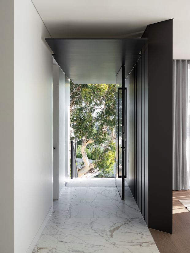 A narrow contemporary hallway with a dramatic black ceiling and full-height black built-in wall or partition on the right. White walls and pale veined stone flooring create a high-contrast, gallery-like passage leading to a glazed opening overlooking trees. A partially visible adjoining room has timber flooring and sheer curtains.