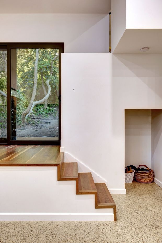 Interior architectural photograph showing a compact split-level circulation area with a stepped timber staircase, polished terrazzo flooring, white plaster walls, and a dark timber-framed glazed door or window overlooking trees. Built-in open storage is visible beneath the upper landing.