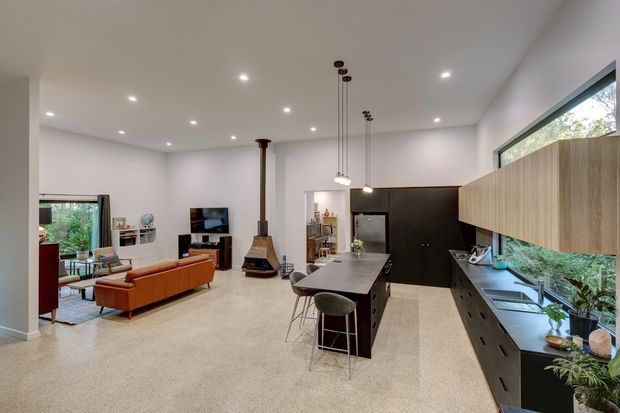 Open-plan contemporary living, dining and kitchen interior with polished concrete flooring, white walls and ceiling, a dark kitchen island and cabinetry, light timber overhead joinery, extensive horizontal glazing, and a suspended fireplace dividing the living area from the kitchen. The room is bright and spacious with integrated recessed lighting and pendant lights.