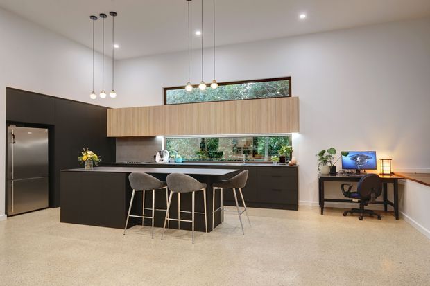 A bright, contemporary open-plan kitchen with a large black island and three bar stools, dark lower cabinetry, warm timber overhead cabinetry, stainless-steel appliances, and a long horizontal window splashback. Pendant lights hang above the island, while a compact home office desk is positioned along the right wall. The room has pale walls, a light polished floor, and a high ceiling.