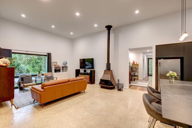 Bright open-plan living area with polished concrete flooring, white walls, recessed ceiling lights, and large windows overlooking greenery. A tan leather sofa faces a television and a freestanding wood-burning fireplace, while a contemporary kitchen island with upholstered stools occupies the foreground. The room connects to a hallway or additional living space through a wide opening.
