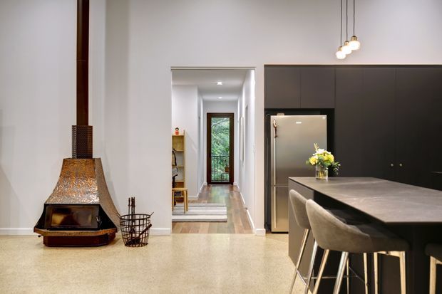 Contemporary open-plan kitchen and dining area with a dramatic freestanding fireplace, dark full-height kitchen cabinetry, stainless-steel refrigerator, stone island with upholstered bar stools, and a polished light terrazzo floor. A hallway extends toward a glazed door and greenery beyond.