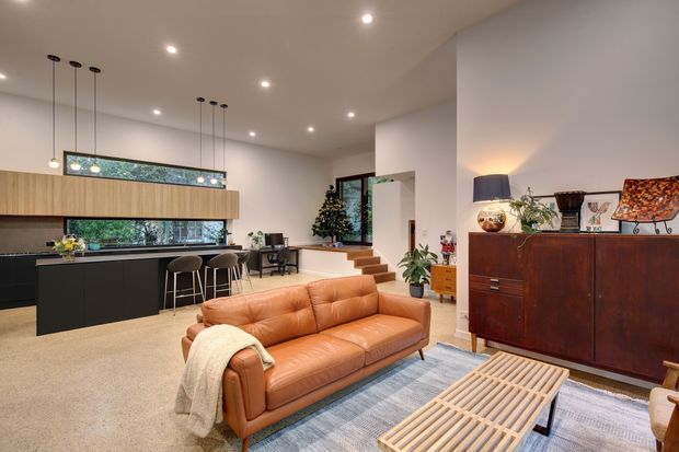 Open-plan contemporary living and kitchen area with a warm mid-century-inspired interior. The room features a black kitchen with timber cabinetry, a long island or peninsula, pendant lighting, a tan leather sofa, patterned rug, timber slatted coffee table, and a dark timber sideboard. A small set of steps leads toward a raised area with extensive glazing and greenery visible outside.