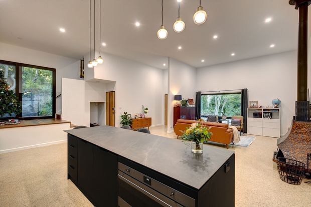 Open-plan contemporary living space with a large dark kitchen island in the foreground, integrated appliances and a pale stone benchtop. The room includes a lounge area with a rust-coloured sofa, white built-in media cabinetry, expansive windows overlooking greenery, pendant and recessed lighting, and a tall black freestanding wood heater.