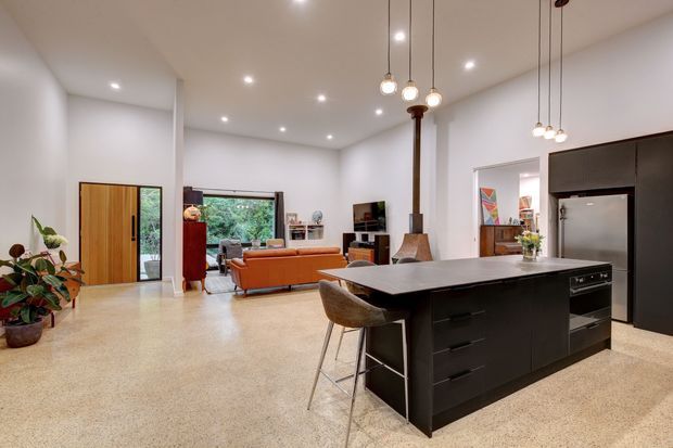 Open-plan contemporary interior combining a black kitchen island with integrated appliances and pendant lighting in the foreground, an adjacent living area with an orange sofa, and polished light terrazzo flooring. White walls, recessed ceiling lights, timber-framed glazing, indoor plants, and a compact freestanding fireplace contribute to the bright, modern space.