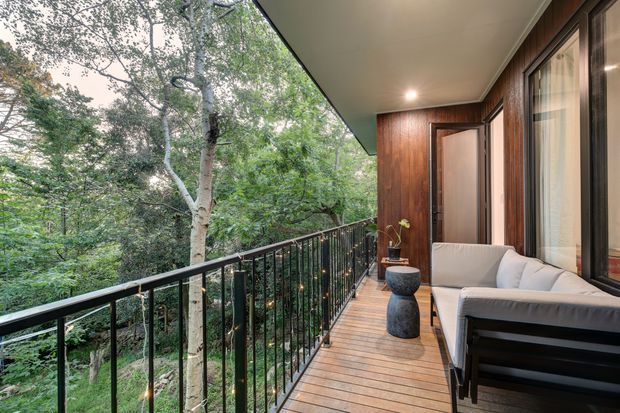 A narrow elevated timber deck or balcony runs alongside a modern home, overlooking dense leafy bushland. The space features dark timber wall cladding, expansive glazed doors and windows, a slim black metal balustrade, recessed ceiling lighting, and an outdoor lounge setting with a light upholstered sofa and small sculptural side tables.