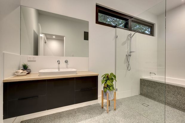 Contemporary bathroom with a double vanity featuring dark fixed cabinetry and a light timber benchtop, a large wall mirror, frameless glass shower enclosure, wall-mounted shower fixtures, high horizontal window, and textured grey floor and lower wall finishes. A small plant adds a natural accent.