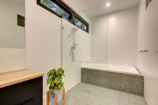 Contemporary bathroom with a large walk-in shower, freestanding-style tiled bath enclosure, frameless glass screen, wall-mounted fixtures, and a speckled terrazzo-like floor and bath surround. High horizontal timber-framed windows bring in natural light, while a dark vanity with a timber benchtop and a potted plant add contrast.