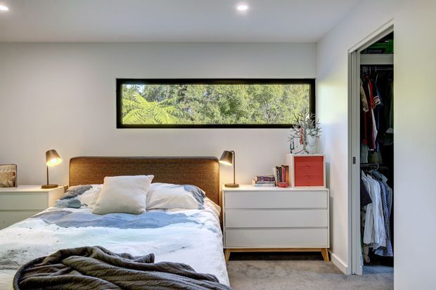 A bright, compact contemporary bedroom with a double bed, woven timber headboard, white bedding, matching white bedside and drawer furniture, and a narrow horizontal black-framed window overlooking lush greenery. An open built-in wardrobe is visible on the right, with recessed ceiling lighting and soft neutral finishes.