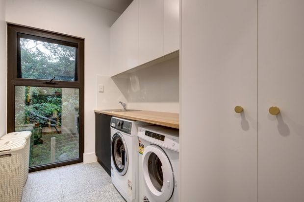 Compact, bright laundry room with stacked-height white cabinetry, timber-look benchtop, two front-loading washing machines, a stainless sink, and a tall dark-framed window overlooking greenery. The room has a light speckled floor and clean contemporary built-in storage.