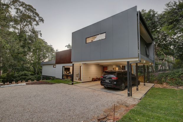 Exterior view of a contemporary two-storey house set among mature trees, with a dark grey upper volume cantilevered over an open carport and covered entry. The lower level combines pale cladding, glazing and warm timber-toned soffit lining, with a gravel driveway and landscaped sloping site.