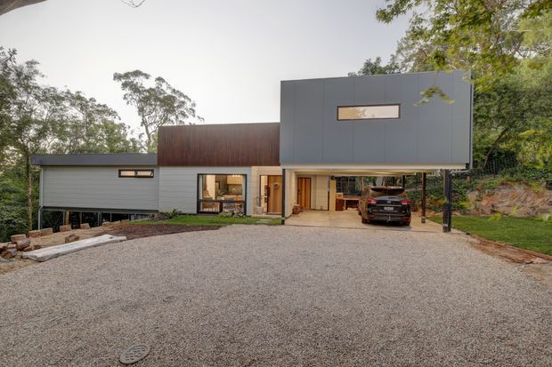Exterior view of a contemporary multi-level house set among trees, featuring dark grey cladding, a projecting upper volume, timber-toned wall panels, narrow horizontal windows, and a covered carport over a broad gravel driveway.
