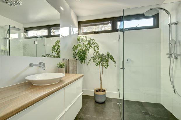Bright contemporary bathroom with a large frameless glass walk-in shower, wall-mounted chrome shower fittings, white wall tiles, dark grey floor tiles, and a floating white vanity with a light timber benchtop and vessel basin. A large mirror, clerestory windows, and a potted indoor tree add light and greenery.