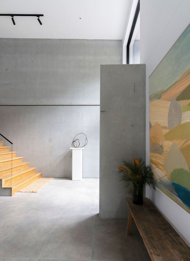A minimalist interior entry or circulation space with a polished concrete floor and smooth concrete walls. A timber staircase rises at the left, while a freestanding concrete partition separates the entry from the adjacent area. A narrow rustic timber console sits beneath a large colourful abstract artwork, with a simple plant arrangement and sculptural object adding restrained decoration. Black track lighting and tall glazing contribute to the bright, contemporary gallery-like atmosphere.