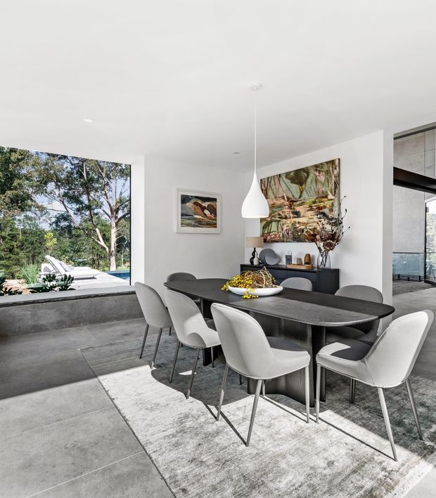 Contemporary open-plan dining area with a dark rectangular dining table surrounded by upholstered light-grey chairs. The room has a white ceiling and walls, large grey concrete-look floor tiles, a pale area rug, minimalist pendant lighting, and a dark sideboard with artwork and decorative objects. Floor-to-ceiling glazing opens the space to leafy outdoor surroundings and a swimming pool area.