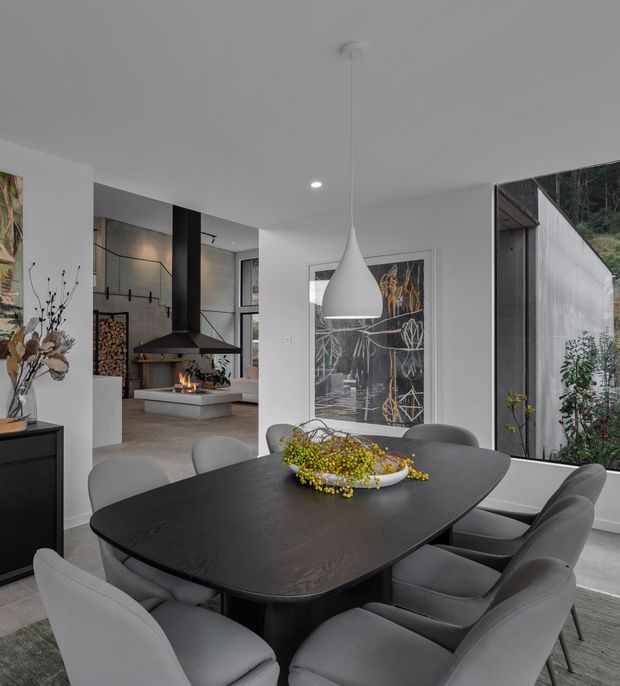 Contemporary open-plan dining area with a dark sculptural dining table and upholstered grey chairs beneath a white pendant light. The space opens toward a modern kitchen and lounge, with a linear fireplace, stacked firewood, large artwork, pale flooring, and expansive glazing overlooking greenery.