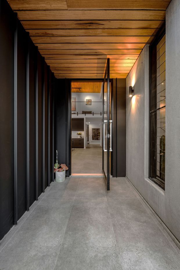 A narrow covered entry passage leading into a contemporary home. Large grey concrete-look floor tiles run between dark vertical metal fins on the left and a pale rendered wall with a gridded window on the right. A warm timber-lined ceiling, black-framed glazed entry door, and wall-mounted lighting create a dramatic, sheltered approach. The interior foyer and a mezzanine-like upper opening are visible beyond the doorway.