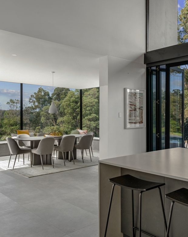 A bright, minimalist open-plan interior showing a dining area adjoining a kitchen peninsula. The dining space has a large rectangular table with upholstered grey chairs, a sculptural pendant light, and floor-to-ceiling glazing overlooking leafy vegetation. In the foreground, a pale kitchen island or breakfast bar has a smooth benchtop with black stools. White walls and ceilings, dark-framed glazing, and large-format grey floor tiles create a clean contemporary setting.