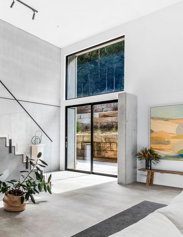 Bright, minimalist double-height bedroom with a partially visible bed in the foreground, polished concrete surfaces, full-height black-framed glazing, and a sculptural open stair along the wall. A concrete structural column, contemporary console, indoor plant, and abstract artwork complete the refined interior. The glazing looks onto a natural, terraced rock landscape.