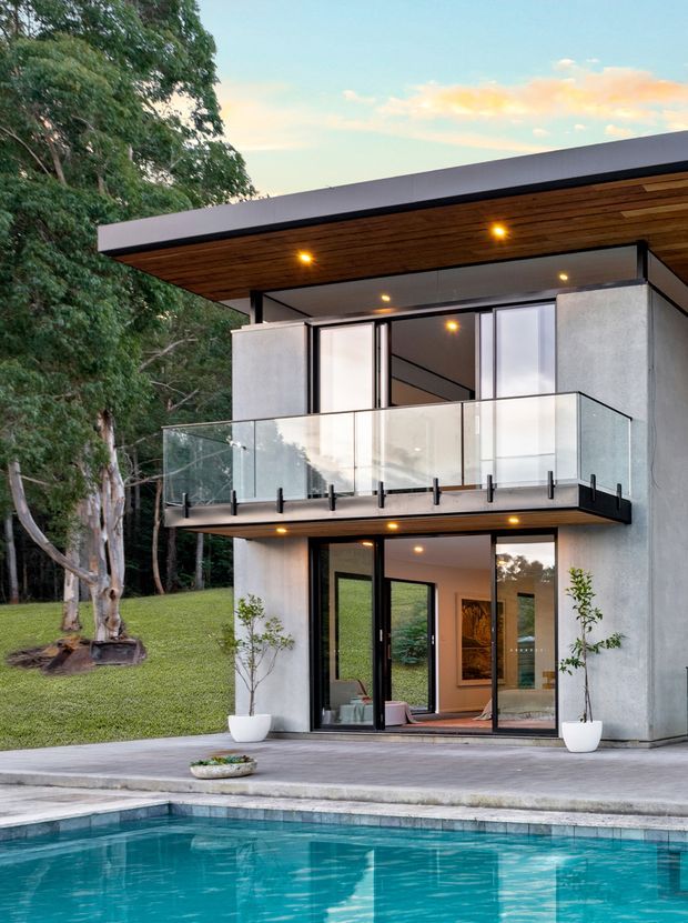 Exterior view of a contemporary two-storey house beside a swimming pool, set within a leafy landscape. The home has a flat roof with deep timber-lined eaves, smooth light-grey rendered walls, extensive dark-framed glazing, and a projecting upper-level balcony with clear glass balustrades. Warm recessed lighting illuminates the timber soffit and balcony edges. Ground-level glazed doors open toward the pool terrace.