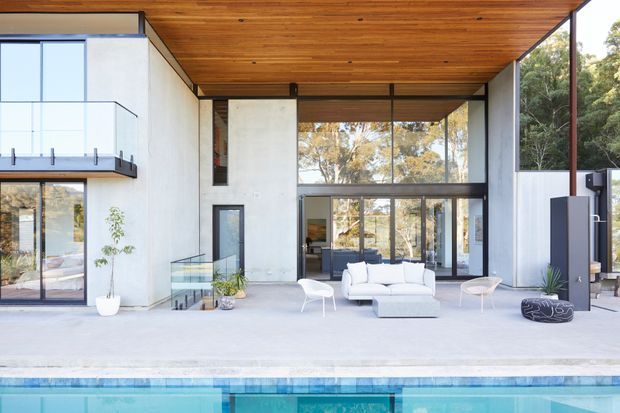 Contemporary modern home exterior viewed from the pool, featuring a broad covered outdoor living area with white lounge furniture, extensive floor-to-ceiling glazing, pale rendered or concrete walls, dark metal-framed doors and windows, and a warm timber-lined soffit. The pool occupies the foreground, while trees are visible beyond the glass and around the building.