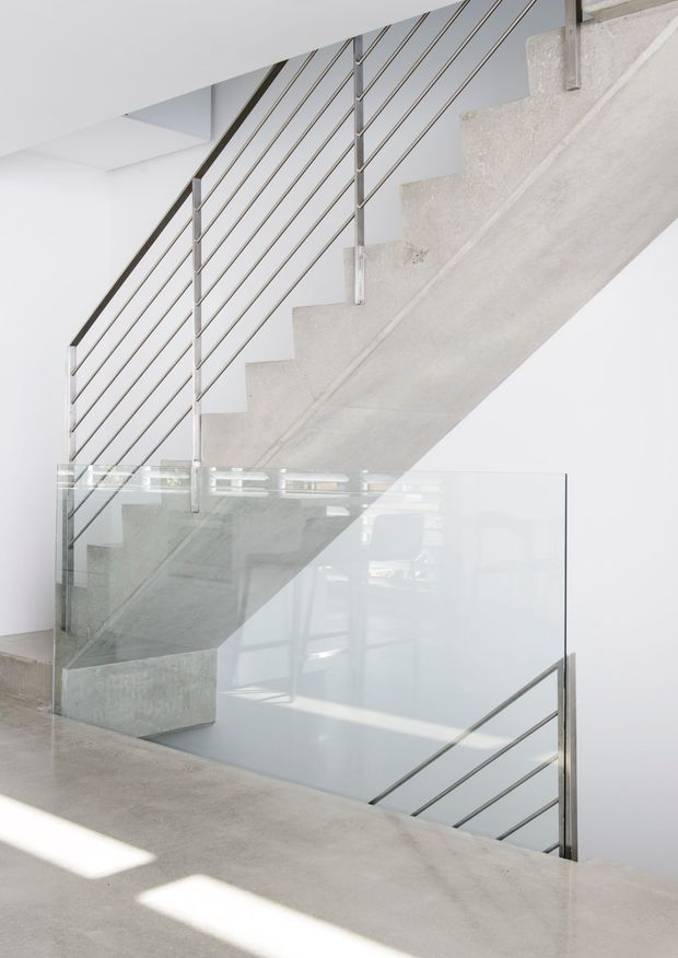 Minimalist interior stairway with a raw concrete staircase, slim horizontal metal balustrades, and a clear glass guardrail around the stairwell. The bright white space has polished or smooth concrete flooring and abundant natural light, creating a clean, contemporary architectural composition.