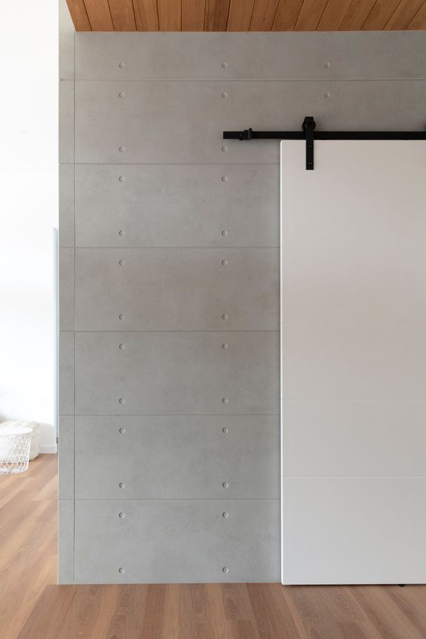 Close-up interior view of a minimalist partition or sliding door assembly. Large smooth grey concrete panels with regularly spaced circular form-tie marks form the main wall, paired with a white sliding door on a black exposed metal barn-door track. A warm timber-lined ceiling and timber flooring add contrast.
