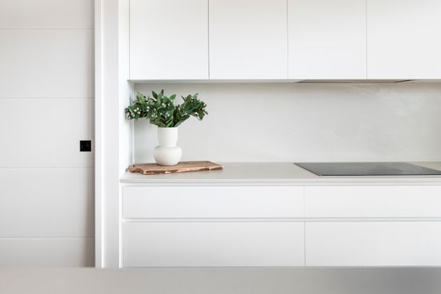 A minimalist white kitchen with flat-panel overhead and base cabinetry, a pale grey benchtop, integrated dark cooktop, and a simple white backsplash. A small potted plant and wooden serving board add subtle natural accents.