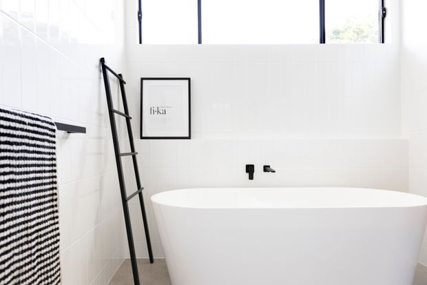 Bright, minimalist bathroom featuring a large freestanding white bathtub, white tiled walls, black tapware, black-framed high windows, and a black leaning towel ladder. A monochrome striped towel and framed artwork add subtle contrast.