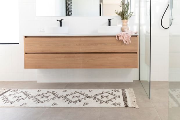 Bright contemporary bathroom featuring a wall-mounted natural timber double vanity with two white basins and matte black tapware, a large mirror, pale tiled walls and floor, and a frameless glass shower enclosure. A patterned runner and small plant add warmth and texture.