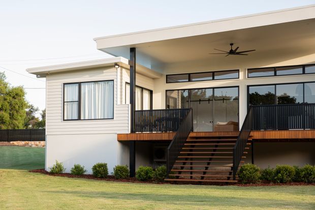 Exterior view of a contemporary single-storey home with a broad flat-roofed covered terrace, pale horizontal cladding, extensive dark-framed glazing, and a raised timber deck accessed by central stairs. Black metal balustrades and landscaped lawn surround the house.