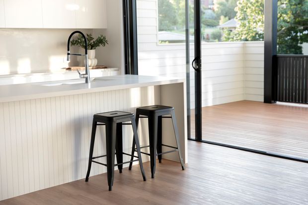 Bright contemporary kitchen with a long white island featuring vertical panel detailing, pale benchtop, white upper cabinetry, and a dark gooseneck tap. Two black metal stools sit at the island. Full-height glazed doors open to a timber deck, bringing greenery and a leafy residential setting into view.