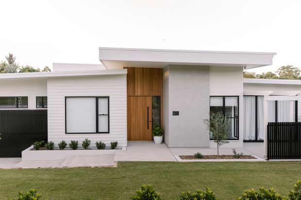 Street-facing view of a low, single-storey modern residence with a flat, slightly projecting roofline, white rendered and horizontal-clad walls, a warm timber entry door, black-framed windows, and a manicured front lawn with simple planted beds. The facade has clean geometric volumes and sheltered outdoor areas.