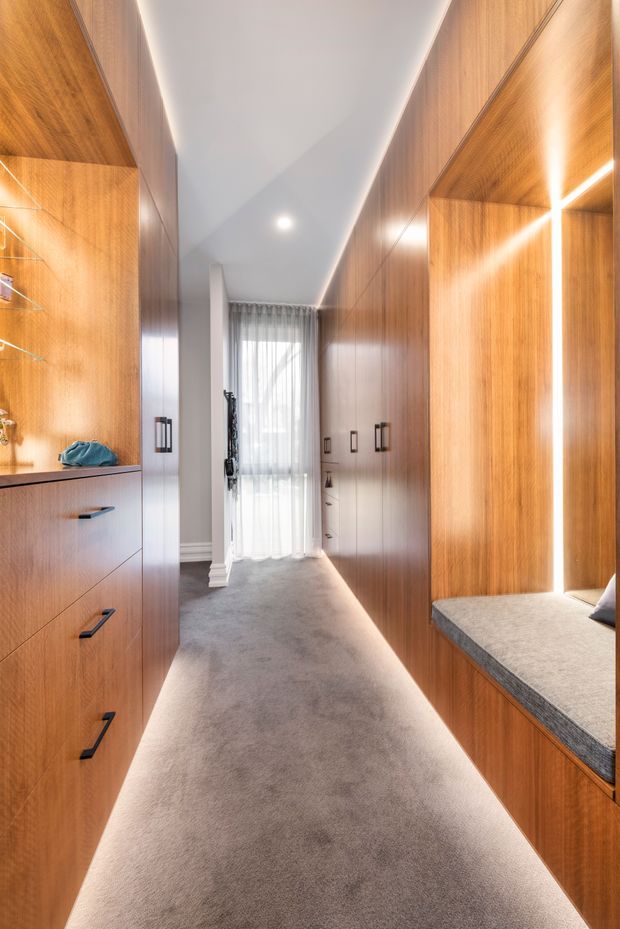 A narrow, refined walk-in wardrobe or dressing corridor lined with extensive warm timber cabinetry on both sides. Built-in drawers, open shelving, hanging storage, integrated lighting, and a cushioned grey bench define the space. Soft carpet, a white ceiling with recessed lighting, and sheer curtains at the far end create a warm, calm, contemporary interior.