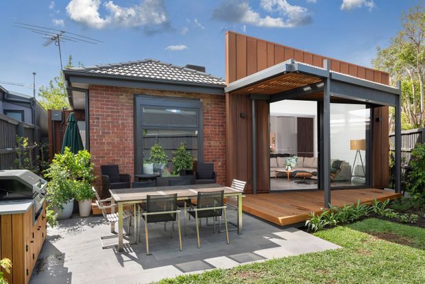 A contemporary residential backyard showing a compact brick dwelling with a dark tiled low-pitched roof and a timber-clad glazed extension. The extension opens onto a raised timber deck beneath a flat-roofed steel-framed canopy, with an outdoor dining setting on a paved patio, planted garden beds, fencing, and a barbecue visible at the side.