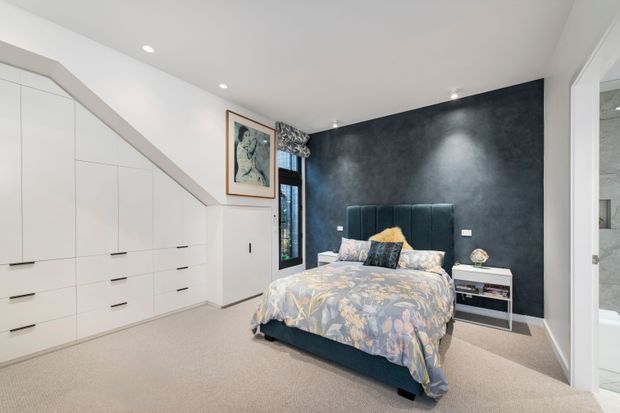 A contemporary bedroom with a large upholstered bed positioned against a dramatic dark charcoal feature wall. The room includes extensive white built-in cabinetry fitted beneath a sloping ceiling, pale carpet flooring, recessed ceiling lights, bedside furniture, framed artwork, and a glimpse of an adjoining tiled bathroom through the doorway on the right.