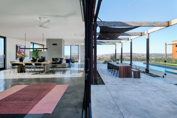 A contemporary open-plan living and dining area opens directly onto a large covered outdoor terrace beside a swimming pool. The interior has expansive black-framed glazing, polished grey concrete flooring, white walls, a timber dining table, and minimalist furnishings. The terrace features a timber pergola, concrete paving, outdoor dining furniture, and views across a sunlit landscape and water.