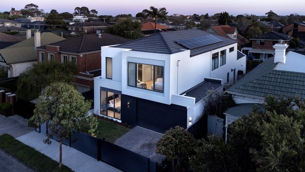 Aerial dusk view of a contemporary two-storey urban residence surrounded by established suburban homes. The house has a crisp white rendered facade, dark vertical cladding and garage door, charcoal pitched roof with an array of solar panels, large windows, and a small front driveway and landscaped garden enclosed by a dark fence.