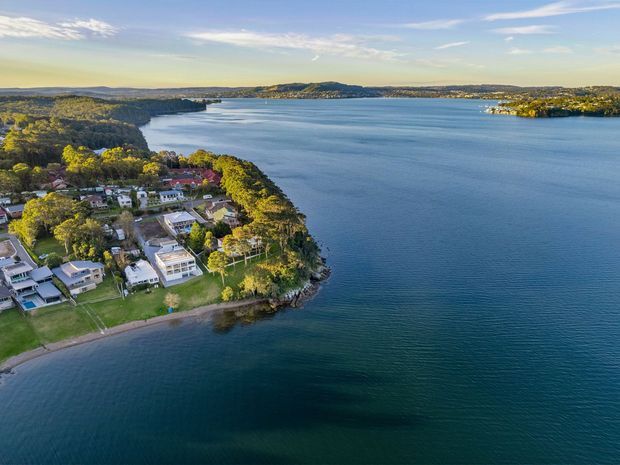 Aerial view of a residential waterfront peninsula surrounded by calm blue water, with detached homes, landscaped lawns, mature trees, and a gently curving shoreline. The image is captured in clear daylight with distant wooded hills and suburban development across the water.