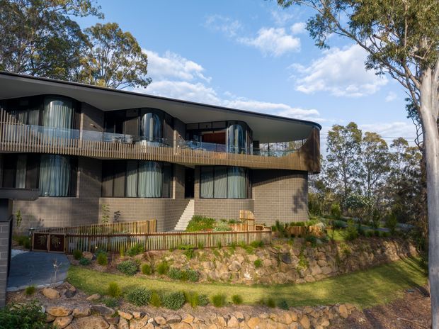 A contemporary two-storey residential building set within a eucalyptus landscape. The dark brick facade has a broad curved roofline and projecting upper-level balcony with timber-look vertical balustrading. Rounded glazed bays and floor-to-ceiling curtains punctuate the facade, while landscaped terraced stone retaining walls and native planting surround the house.