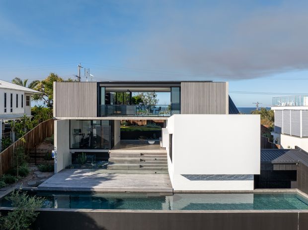 Contemporary multi-level residential house viewed from the rear, featuring a boxy flat-roof form, vertical timber-look cladding, extensive glazing, a stepped terrace, and a long swimming pool in the foreground. The property appears elevated on a sloping suburban coastal site, with ocean visible beyond the house.