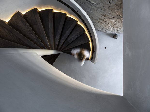 View looking down a sculptural spiral staircase with dark timber wedge-shaped treads, smooth pale plaster or concrete walls, and warm concealed lighting tracing the stair edge. A rough exposed-stone ceiling or wall surface is visible at the upper right, with a blurred figure or object on the stairs. The composition emphasizes the dramatic curved geometry and contrast between dark wood, pale walls, and textured stone.