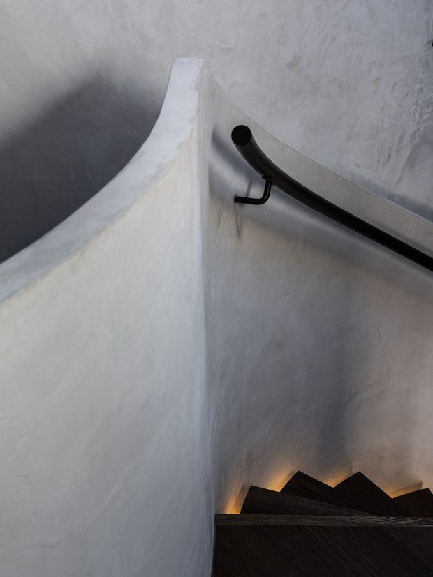 Close-up architectural view of a curved stairway with sculpted white plaster or rendered walls, a dark timber stair flight, and a black curved metal handrail. Warm concealed lighting glows beneath the stair treads, creating a dramatic, sculptural interior.