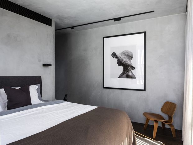 A compact, minimalist bedroom with a low upholstered dark headboard, crisp white bedding, and a dark taupe-brown throw. The room features mottled grey plaster walls, a dark timber floor, recessed linear black ceiling lighting, a framed monochrome portrait, and a sculptural wooden lounge chair beside sheer curtains. The restrained palette and textured finishes create a calm, contemporary atmosphere.