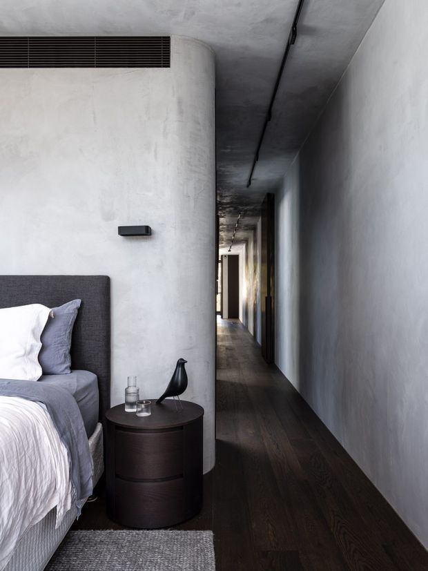 A moody, minimalist bedroom opening onto a long narrow hallway. The room features a dark upholstered bed with grey and white bedding, a cylindrical dark wood bedside table, a black sculptural bird ornament, dark timber flooring, and smooth pale grey plaster walls. A curved partition wall separates the bedroom from the corridor, which is lit by recessed or linear ceiling fixtures.