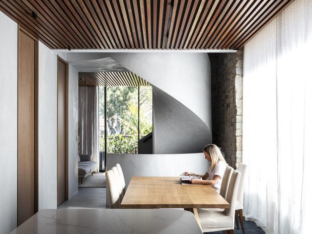 A refined contemporary dining area with a timber dining table and upholstered chairs, framed by a striking slatted timber ceiling. A sculptural curved plaster staircase or architectural partition rises behind the dining zone, with exposed rough stonework along the right wall, full-height sheer curtains, and a glimpse of greenery through glazing. A marble-look benchtop is visible in the foreground, suggesting the dining area is adjacent to a kitchen.