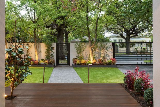 A landscaped residential courtyard viewed from a timber deck, featuring a straight gravel path leading to a black horizontal-slat pedestrian gate and matching vehicle gate. Manicured lawn panels, mature trees, planted garden beds, warm wall lighting, rendered boundary walls, and a partially visible light-coloured house create a refined modern garden setting.